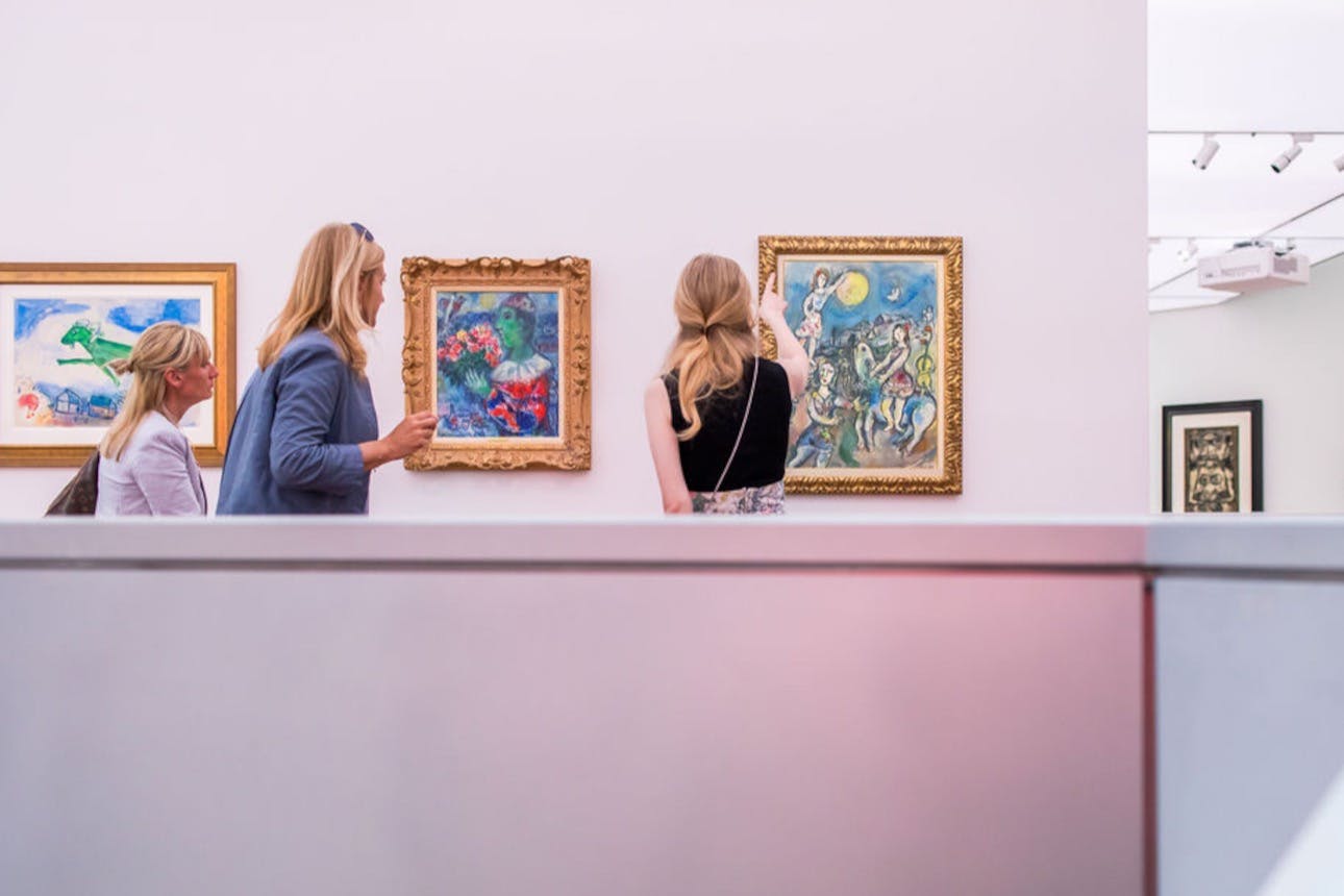 Dvě ženy si v galerii prohlížejí zarámovaná umělecká díla, jedna ukazuje na barevný obraz. V popředí je viditelná přepážka.