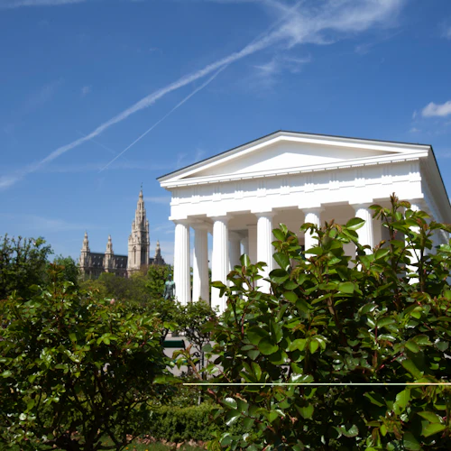 Vit neoklassisk byggnad med pelare omgiven av grönska, avlägsna katedralspiror, under en blå himmel med skumma moln.