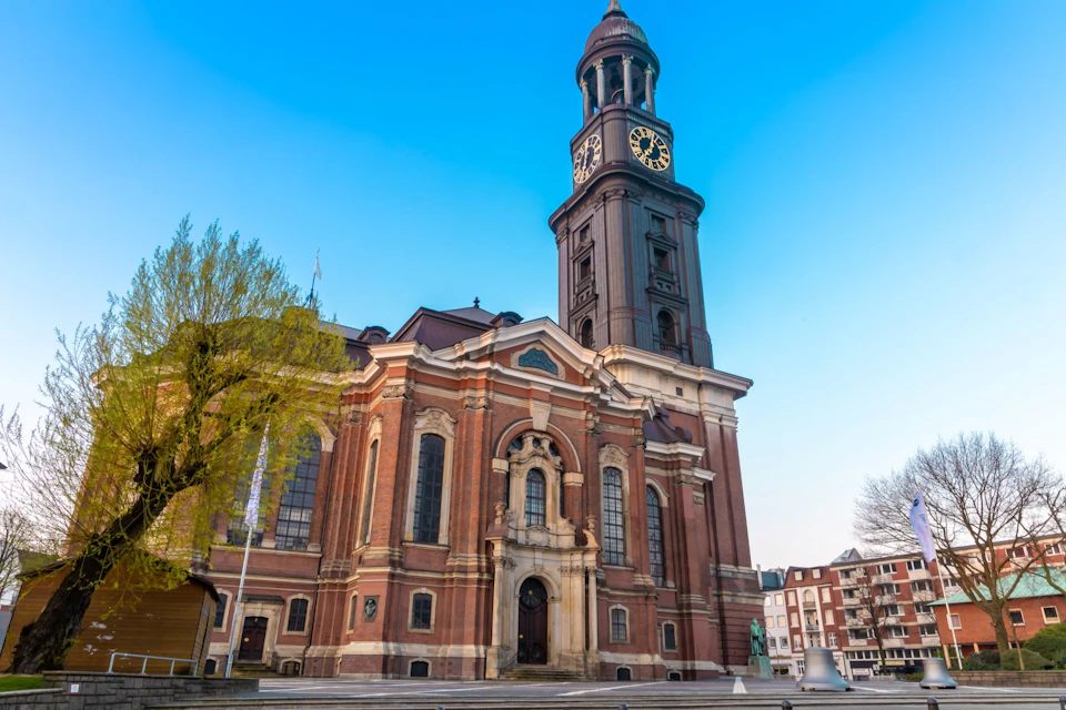 A historic brick church with a tall clock tower against a clear blue sky, surrounded by trees and urban buildings.