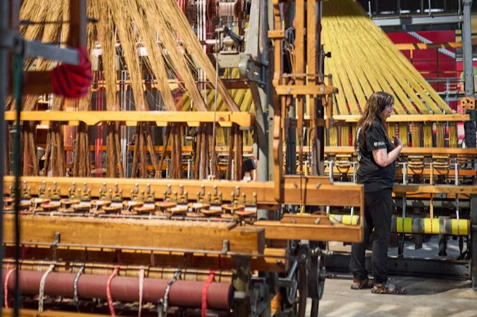 A person standing near a traditional loom in a weaving workshop, with threads and machinery visible.