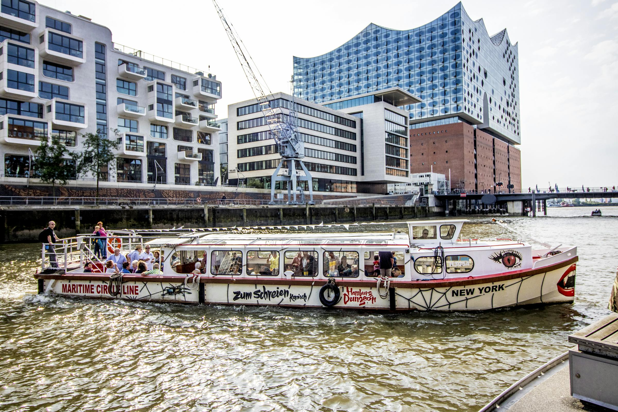 Un bateau d'excursion sur une rivière passe devant des bâtiments modernes et une grue sous un ciel ensoleillé ; des personnes sont visibles à l'intérieur du bateau.