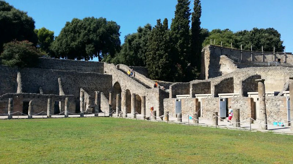 An ancient stone structure with arches and stairs surrounded by grass and trees under a clear blue sky.