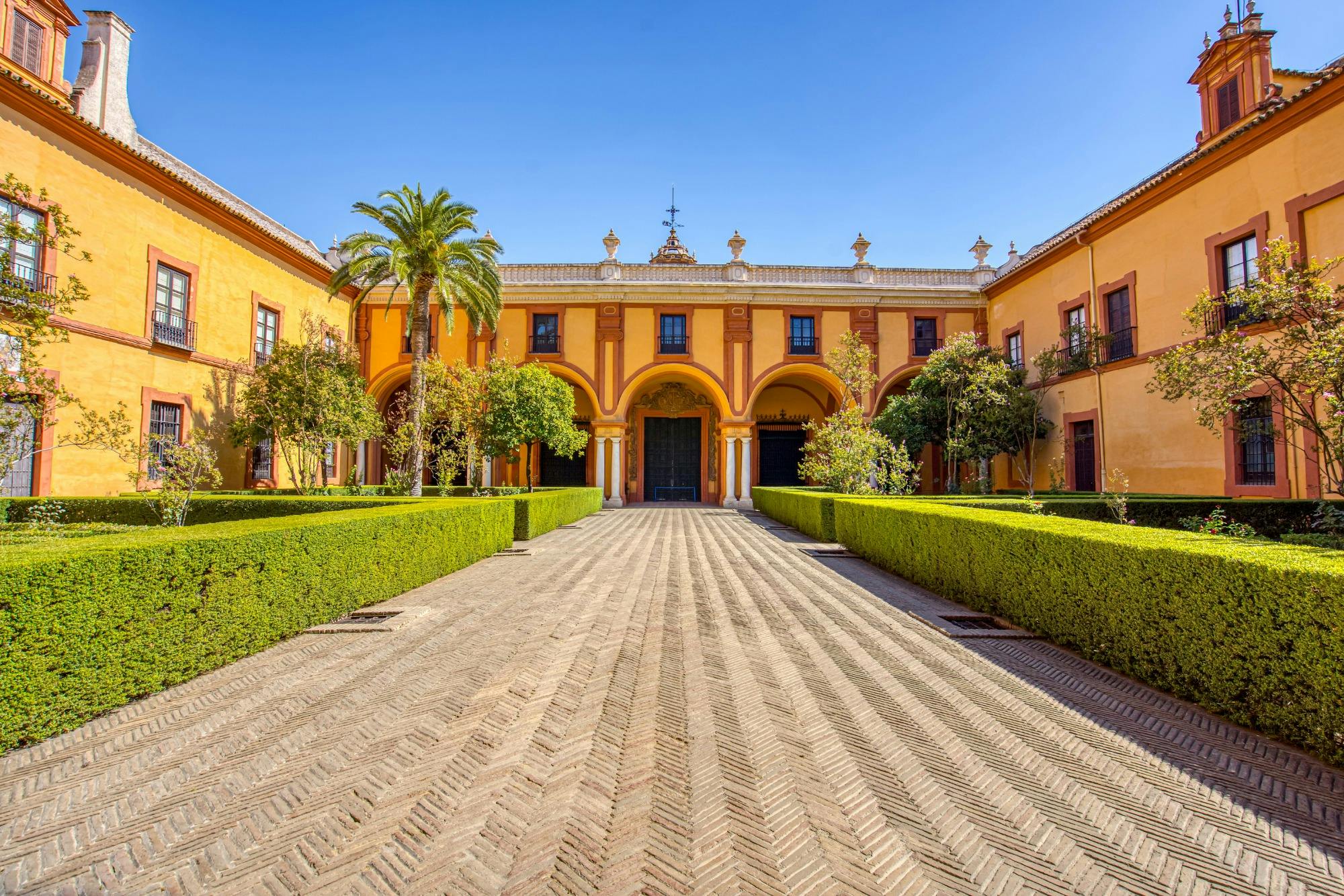 A symmetrical courtyard with a central pathway, bordered by hedges, leading to a grand building under a clear sky.