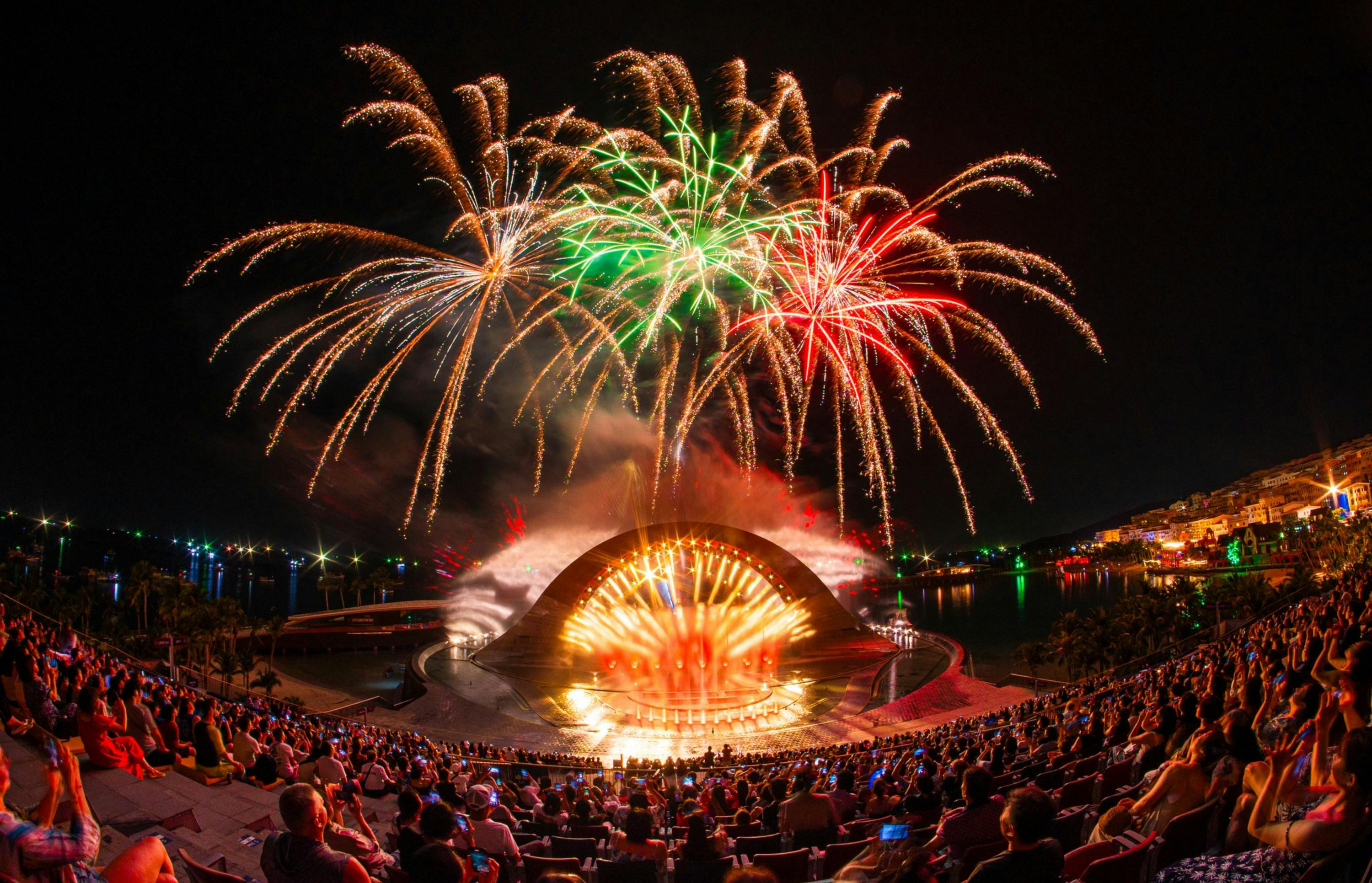 A vibrant fireworks display above an illuminated outdoor stage and amphitheater with a seated audience at night.