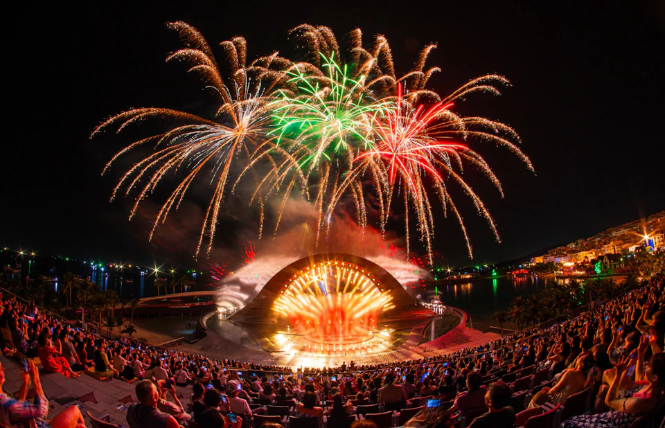 A nighttime outdoor performance with fireworks above a stage and a seated audience in an amphitheater.