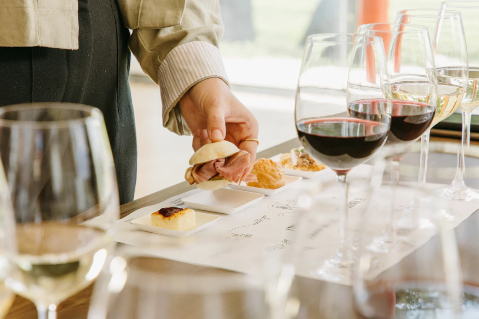 A person holding a small sandwich over a table set with various appetizers and glasses of red wine.