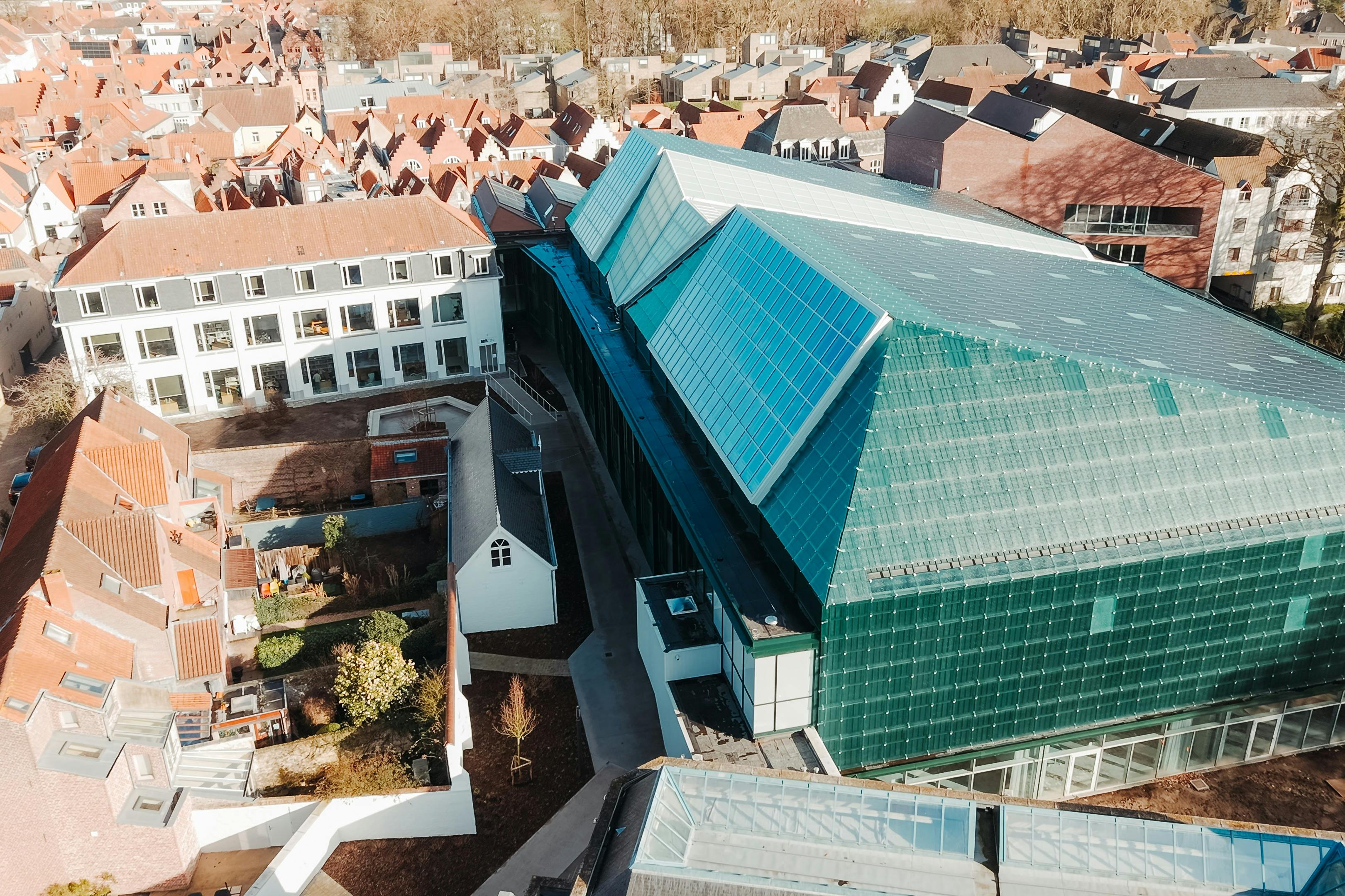 Flygfoto över en modern glasbyggnad intill traditionella tegelkonstruktioner i ett tätt bebyggt område.