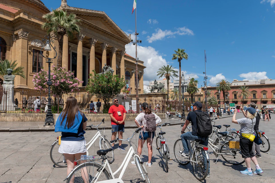 A group of people on bicycles in front of a historic building with columns and palm trees on a sunny day.