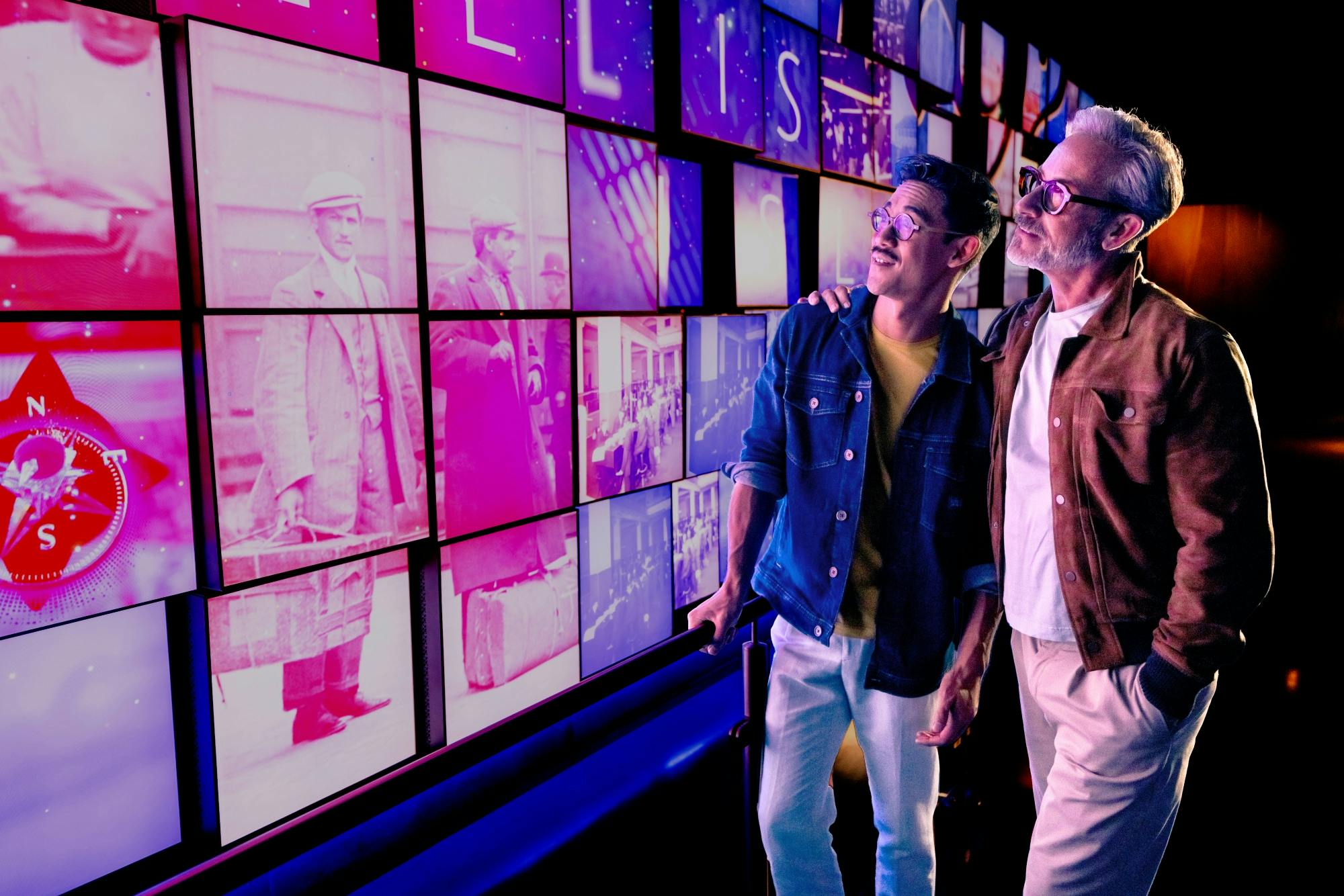 Δύο άνδρες κοιτάζουν έναν τοίχο με φωτισμένες ιστορικές φωτογραφίες και εικόνες, με τον άνδρα στα αριστερά να δείχνει προς την οθόνη.