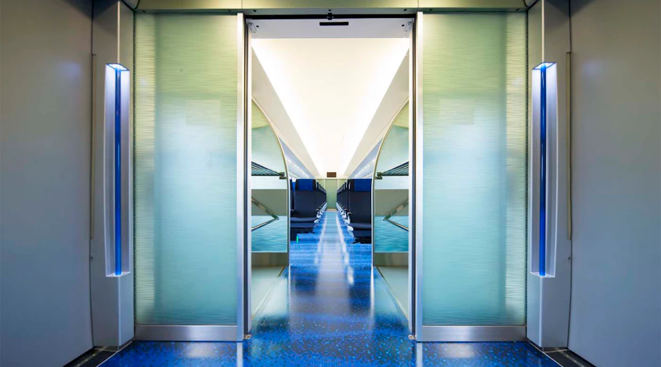 Interior view of a modern train or transit car with blue floors, sleek design, and a bright ceiling, seen through sliding doors.