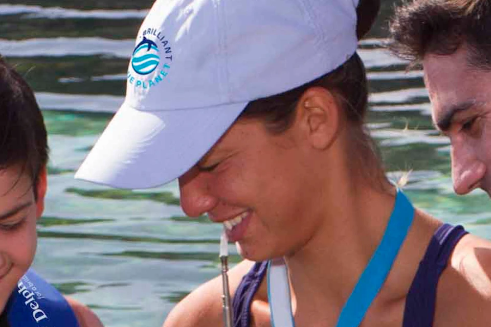 A child and two adults in blue life vests smile while interacting with a dolphin in shallow, clear water.