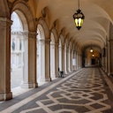 A covered walkway with ornate columns and hanging lanterns, featuring a patterned floor and archways to an open courtyard.