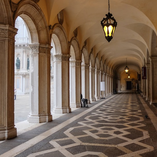 A covered walkway with ornate columns and hanging lanterns, featuring a patterned floor and archways to an open courtyard.