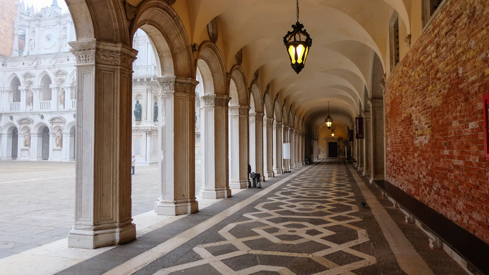 A covered walkway with ornate columns and hanging lanterns, featuring a patterned floor and archways to an open courtyard.