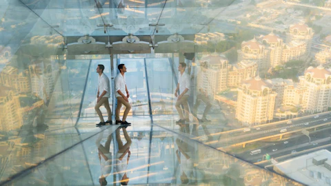 A man in a white shirt and beige pants stands in a glass observation deck with cityscape reflections. "EMAAR" logo at bottom.