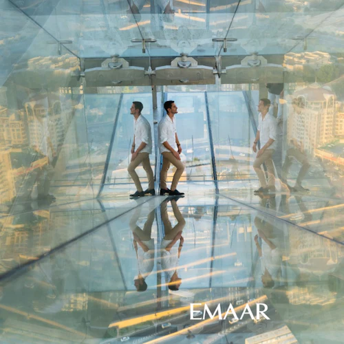 A man in a white shirt and beige pants stands in a glass observation deck with cityscape reflections. "EMAAR" logo at bottom.