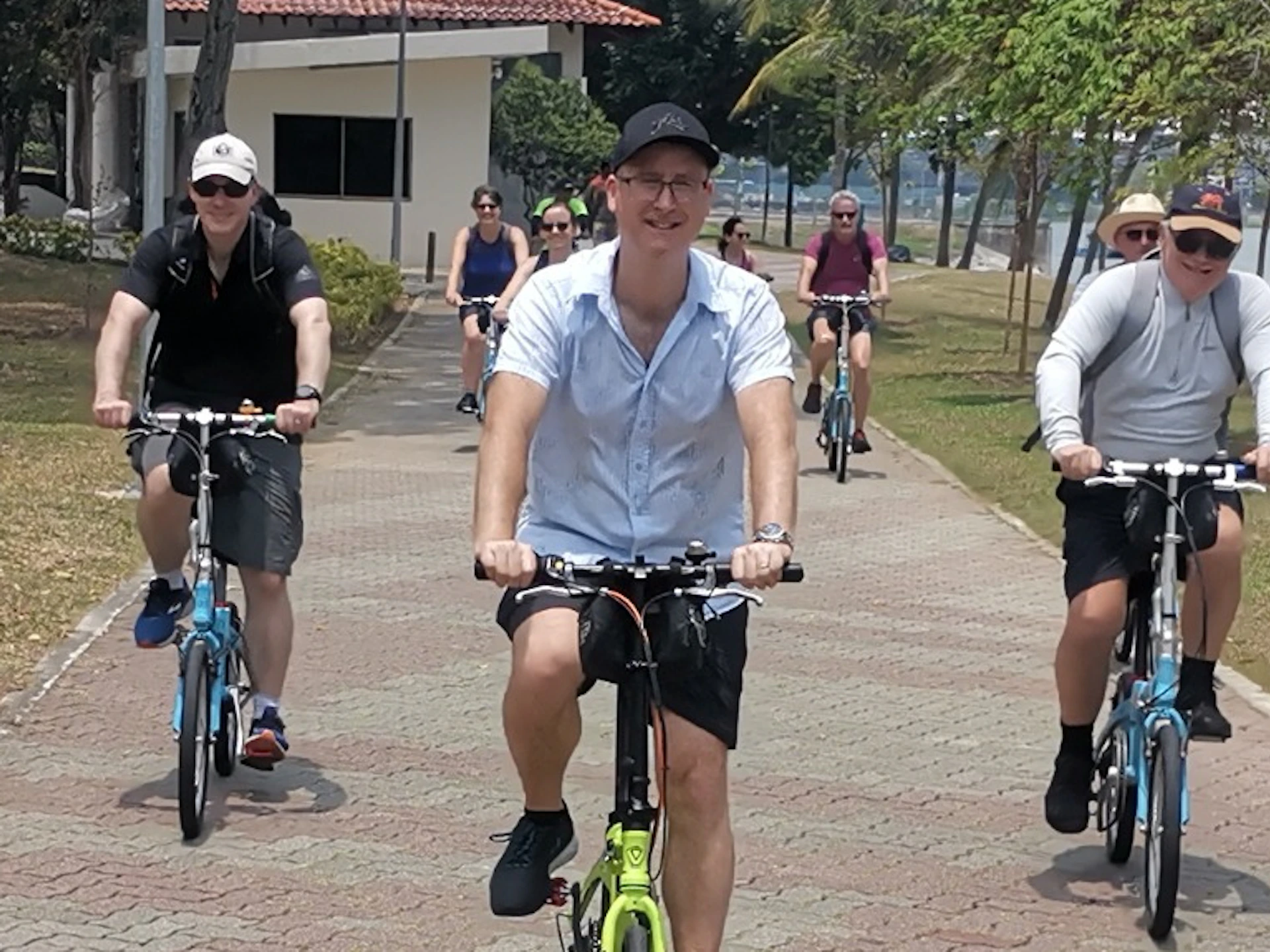 A group of people riding bicycles on a paved path in a park near a body of water, surrounded by trees and greenery.