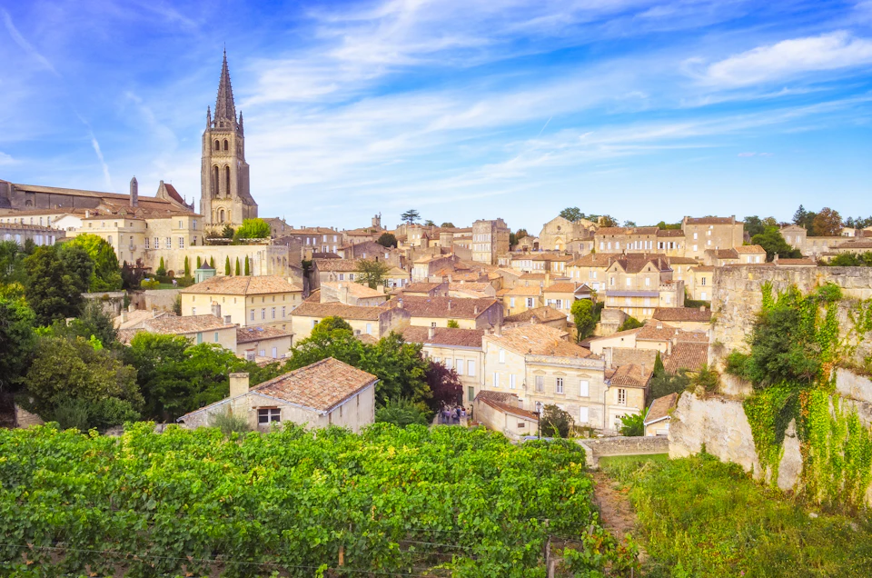 A scenic view of a town with tan-coloured buildings, a church with a tall spire, and green vineyards in the foreground beneath a blue sky.