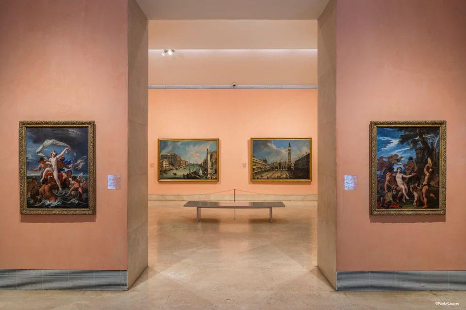 A museum gallery with framed paintings on pink walls and a bench in the center. The floor is polished stone.