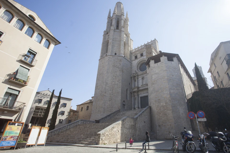 A large stone cathedral with a tall bell tower. A child and two adults stand near the steps with a bicycle on the right.