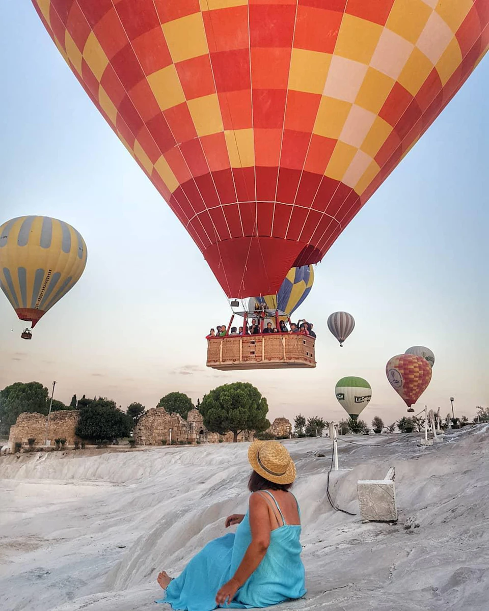 A woman in a blue dress and straw hat watches hot air balloons of various colors lift into the sky over a rocky landscape.