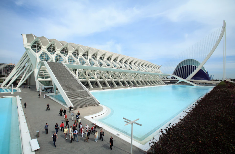 Modern architectural building with a large blue pool and a group of people walking on a pathway beside it under a blue sky.