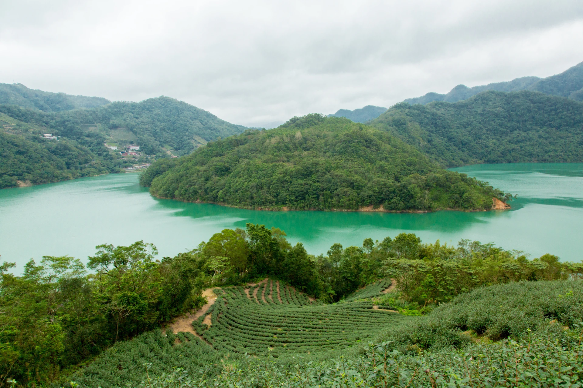 A lush, green island surrounded by a turquoise lake, with terraced fields and distant mountains under a cloudy sky.