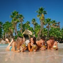 Quattro persone sdraiate nell'acqua bassa di una spiaggia tropicale, che bevono da noci di cocco e ananas, con le palme sullo sfondo.