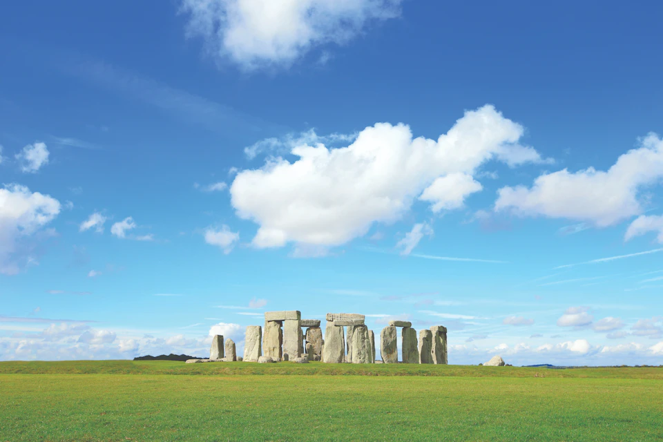 Stonehenge under a blue sky with scattered clouds, surrounded by green grass.