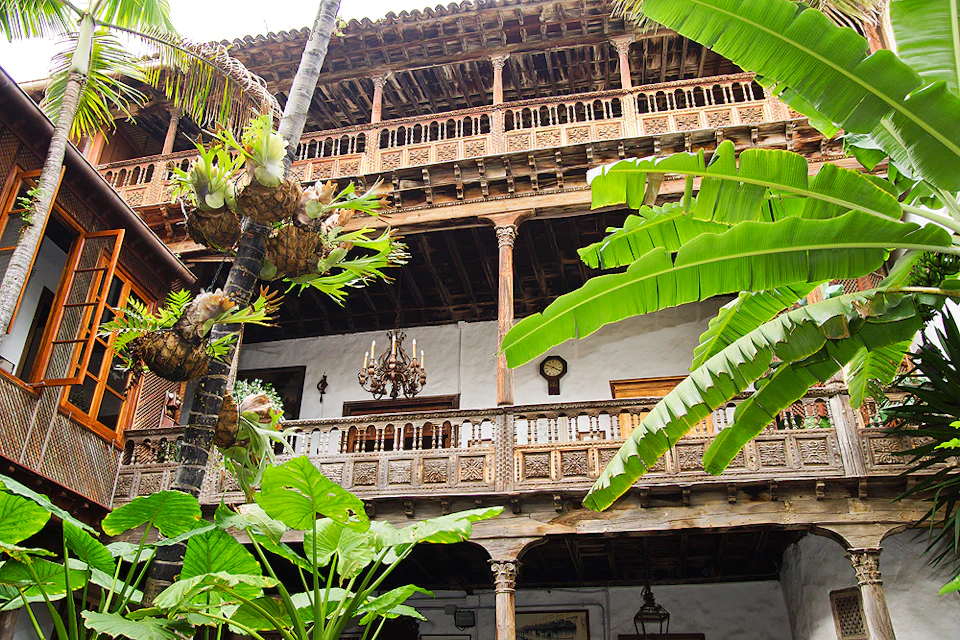 Three-story building with ornate wooden balconies and lush tropical plants in the foreground. A chandelier hangs on the second floor.