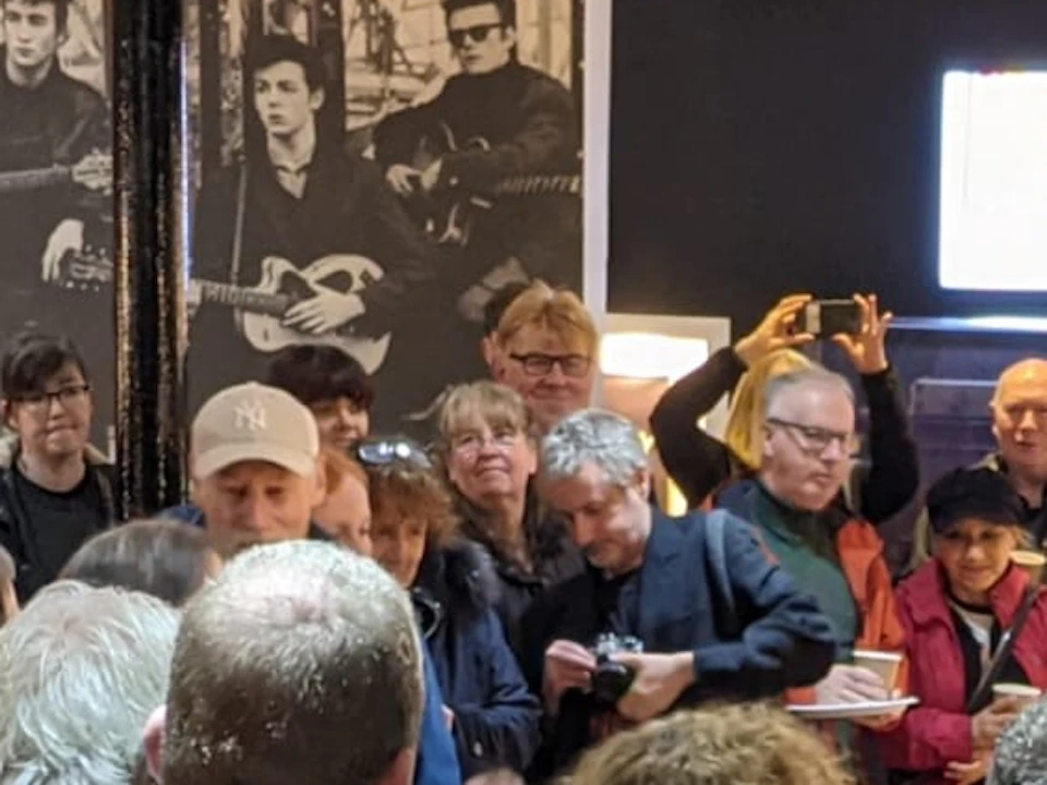 A group of people gathered in a room, some holding records, with black-and-white photos and a lit display screen in the background.