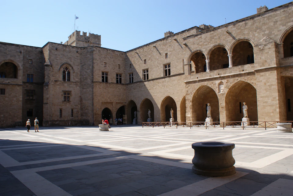 A spacious stone courtyard surrounded by an ancient building with arched walkways and columns. A few statues are visible.