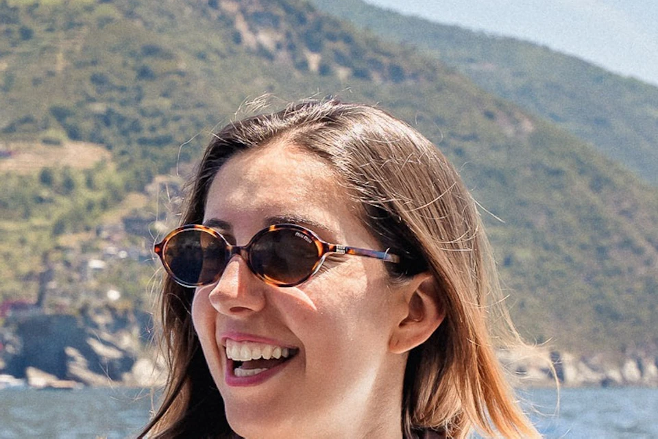 Three women smiling and talking on a boat with a scenic view of hills and water in the background on a sunny day.