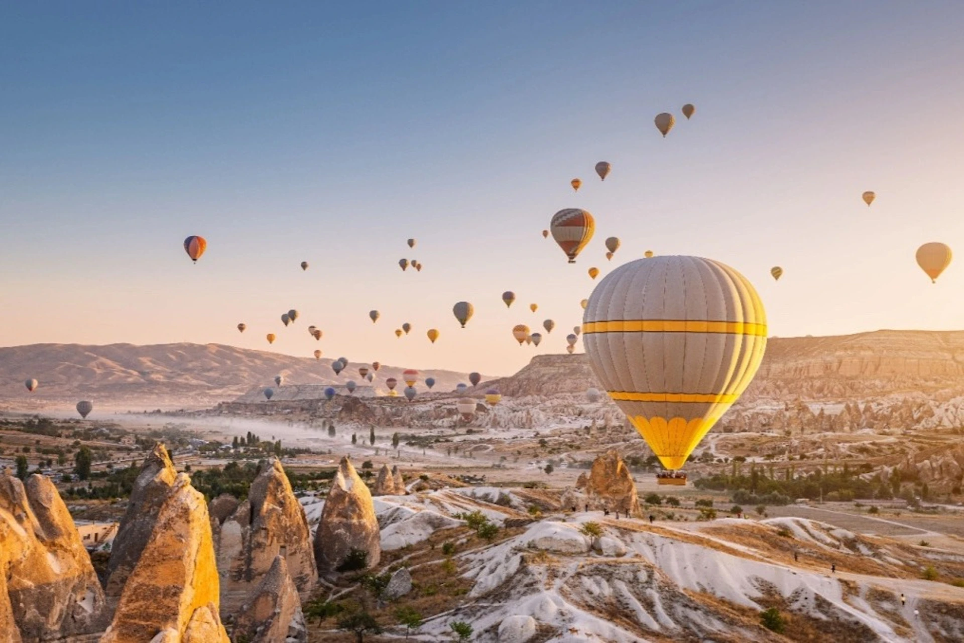Numerous hot air balloons float above a rocky, arid landscape during sunrise, with mountains and valleys in the background.