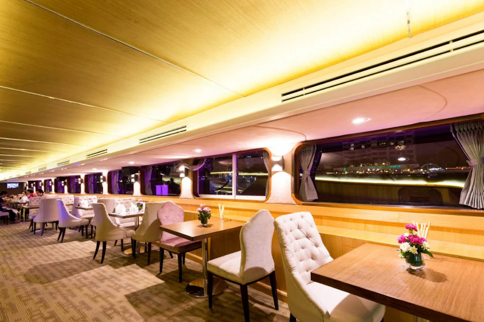 A brightly lit, elegant dining area with upholstered chairs, wooden tables, and large windows offering a nighttime view.