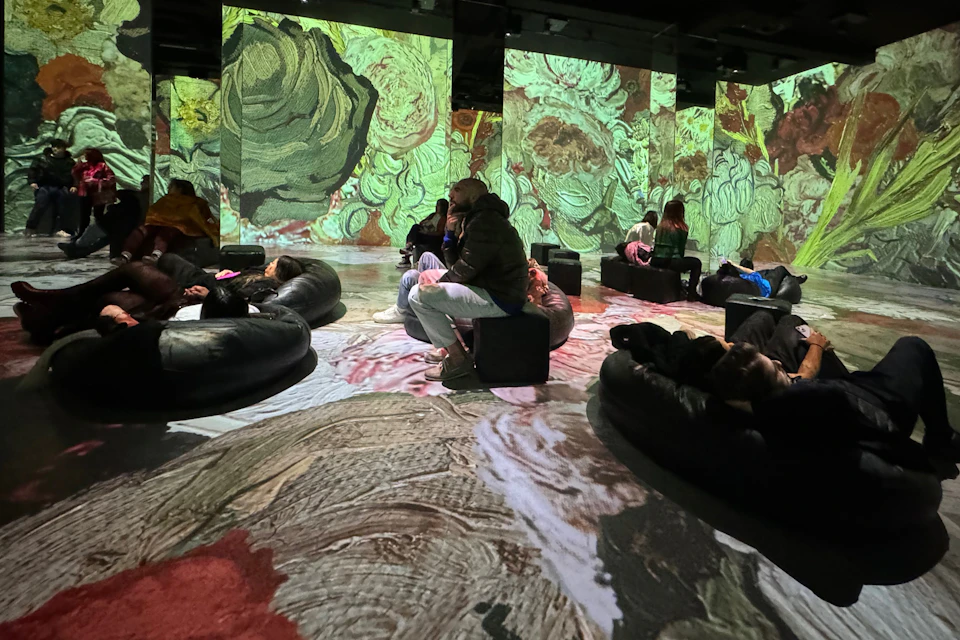 People sitting or reclining on cushions in a room with colorful floral artwork projected on the walls and floor.