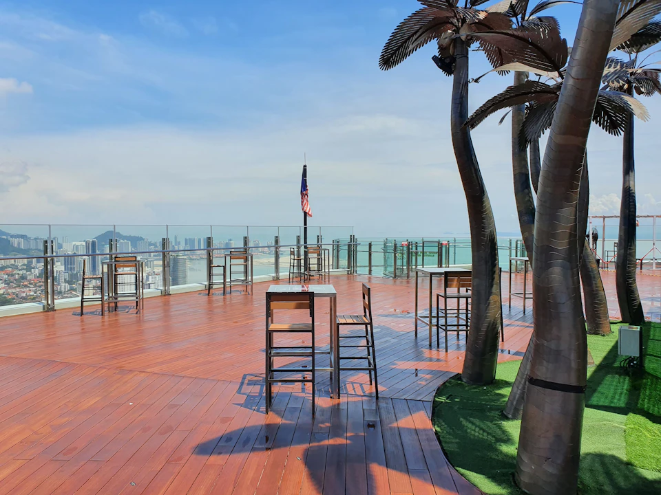 A rooftop deck with wooden flooring, metal chairs and tables, decorative palm trees, and a view of a city skyline.