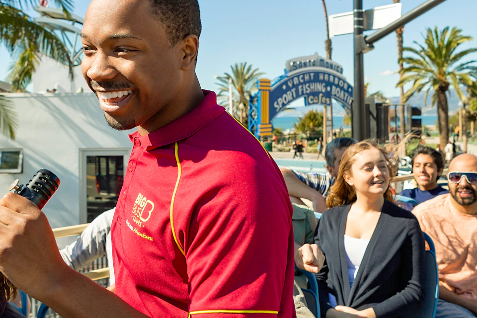 A smiling tour guide with a microphone leads a group of people on an open-top bus tour near a waterfront in sunny weather.