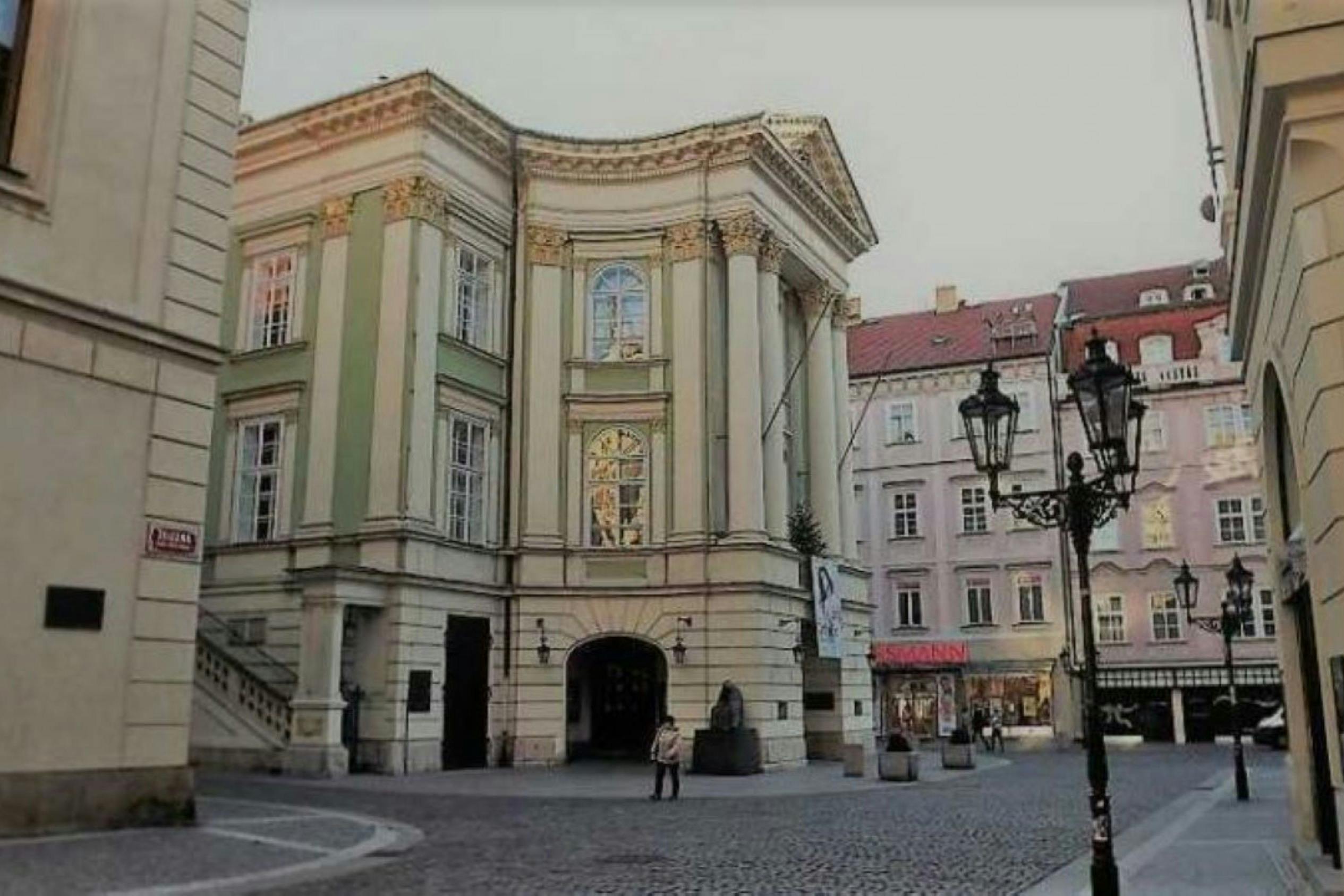 A historic building with large columns beside a cobblestone street, a person standing near a statue, and shops in the background.