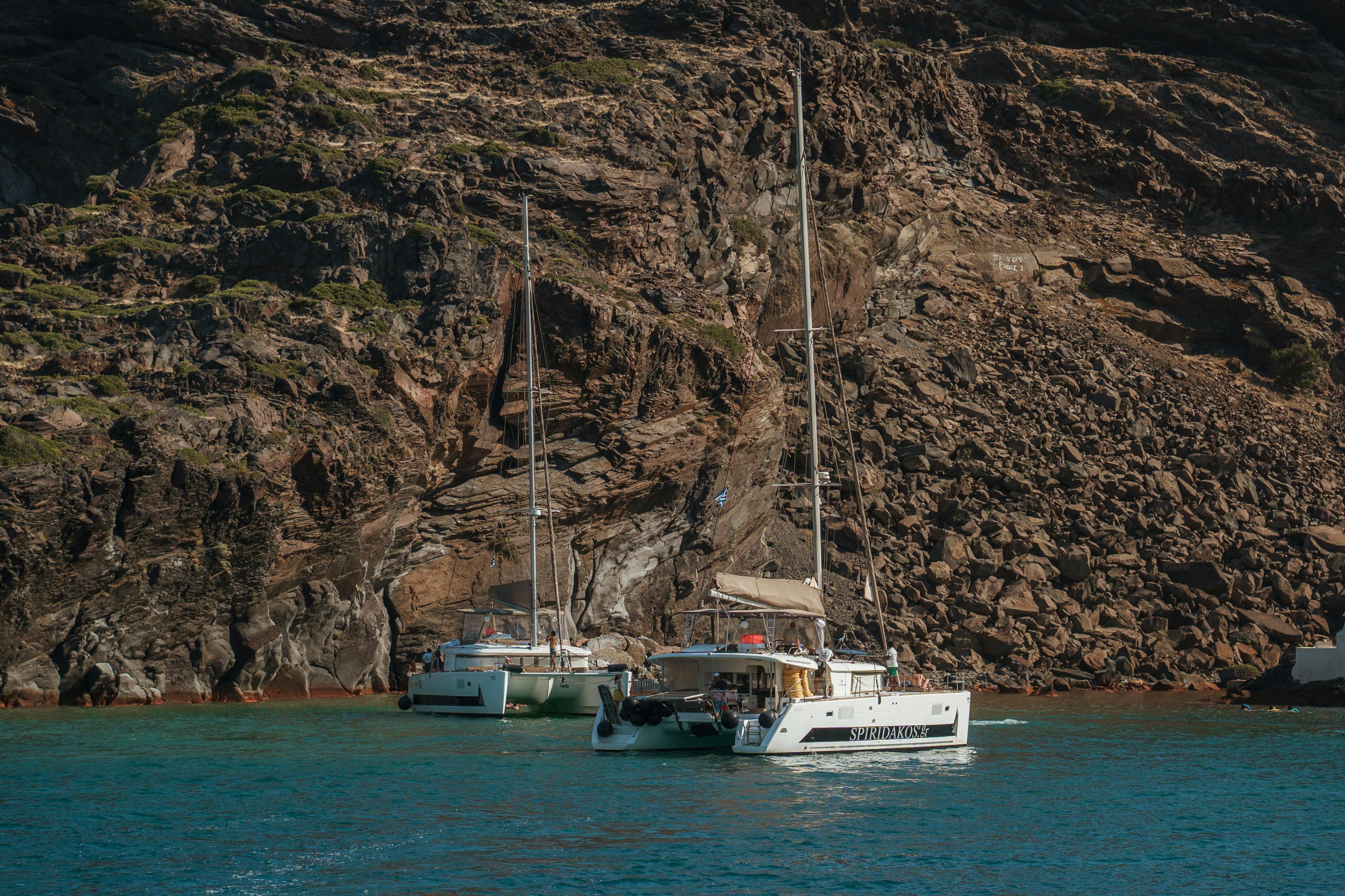 Two sailboats anchored near a rocky shoreline with clear turquoise water and steep cliffs in the background.