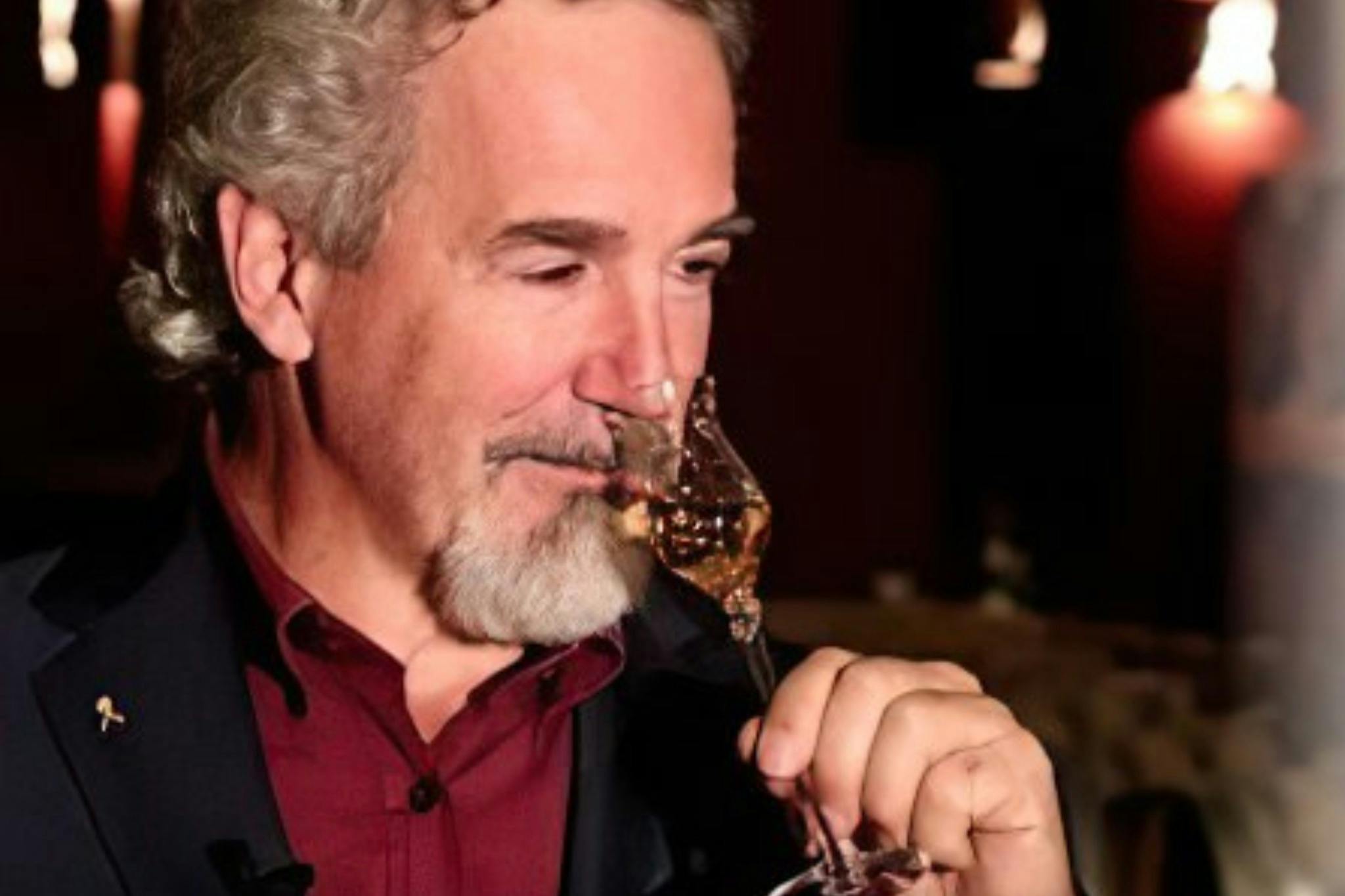 Ένας άντρας με γκρίζα μαλλιά και γένια, φορώντας ένα σκούρο σακάκι και κόκκινο πουκάμισο, χώνει ένα ποτήρι υγρό κεχριμπαριού σε ένα αμυδρά φωτισμένο περιβάλλον.