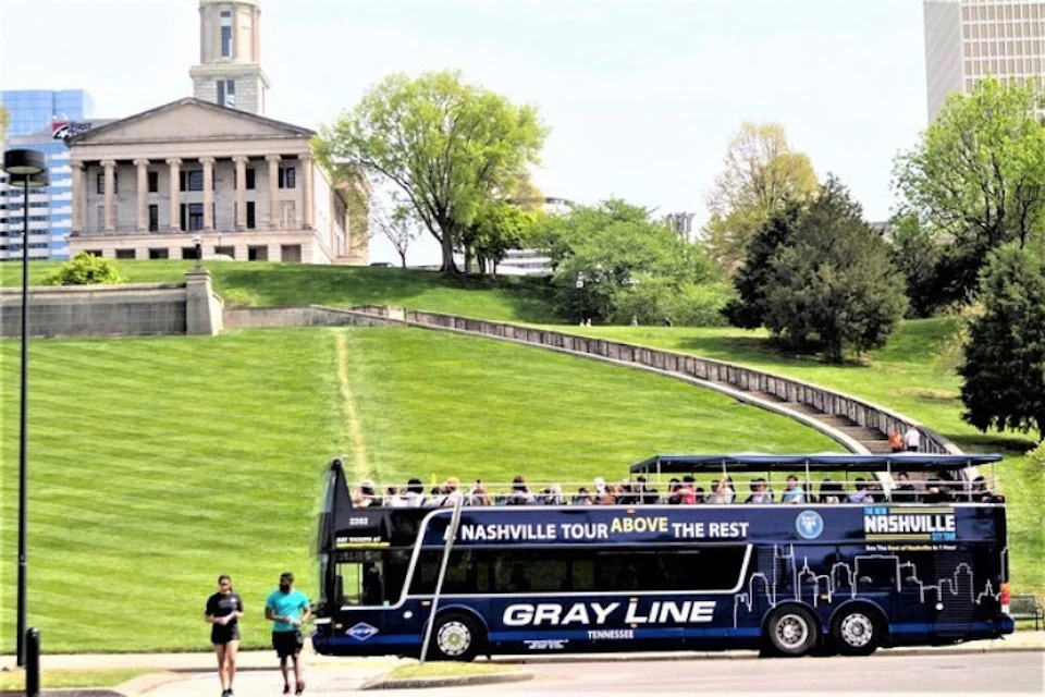 A double-decker tour bus in front of a historic building and manicured lawn with trees. Two people walk nearby.