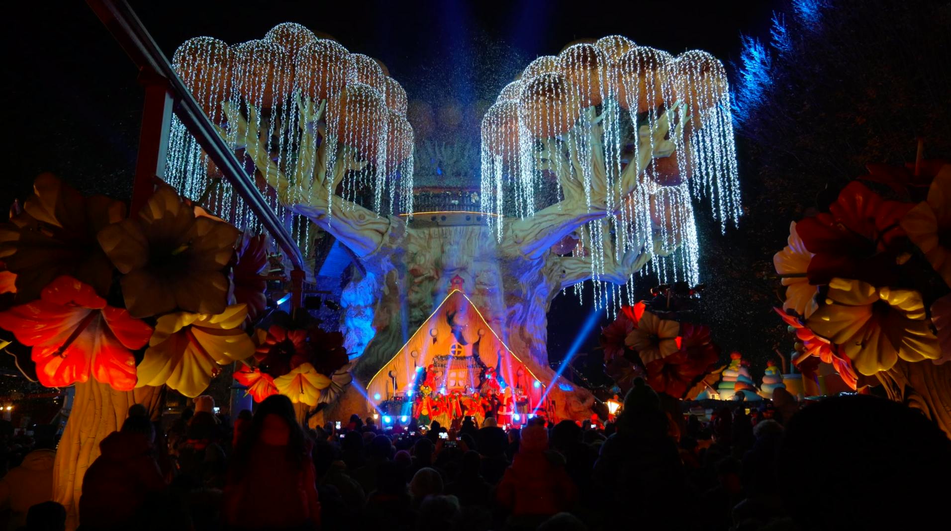 Dav sleduje jevištní představení pod velkými osvětlenými stromy se zavěšenými světly v noci.