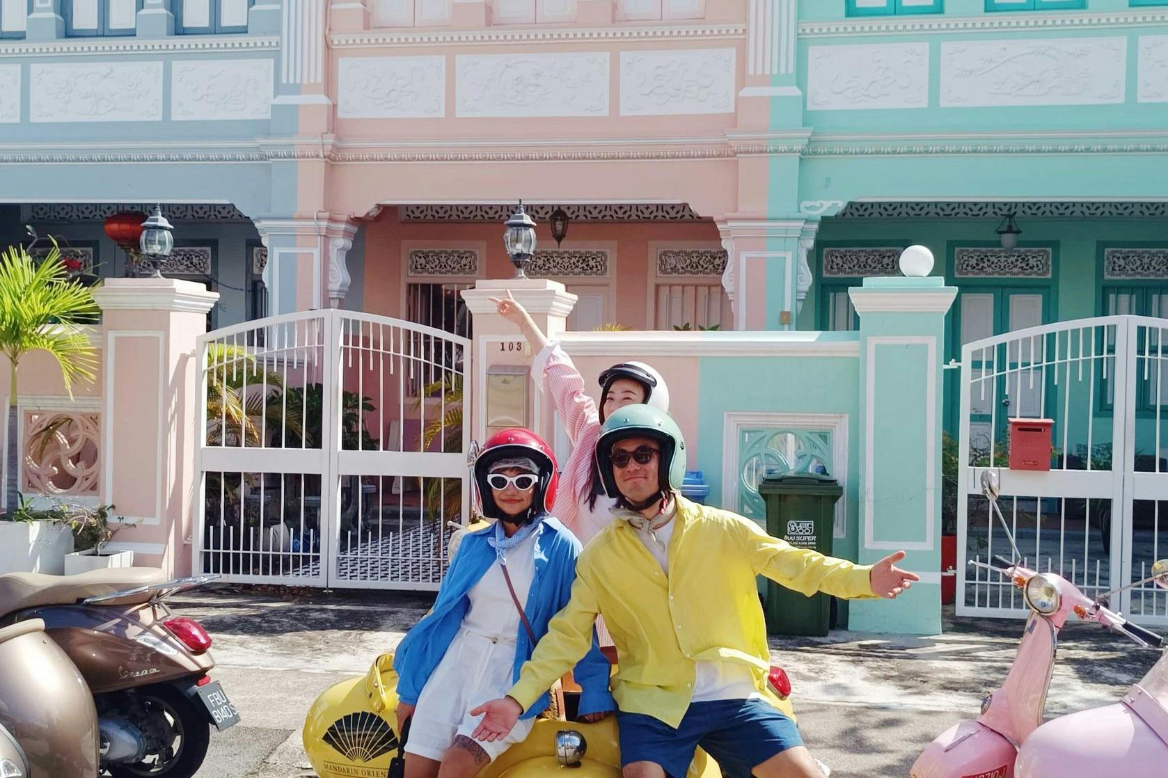 Drei Personen in farbenfrohen Outfits posieren an einem sonnigen Tag auf einem gelben Motorroller vor pastellfarbenen Gebäuden.