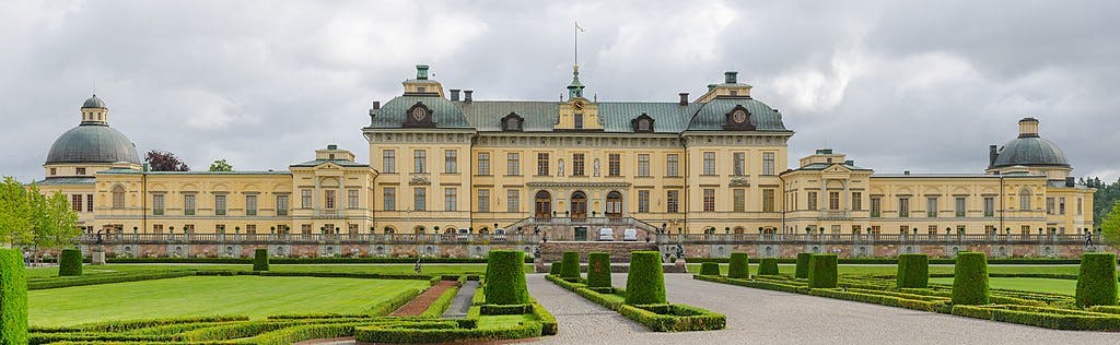 A large yellow palace with green domes and symmetrical gardens in front, featuring neatly trimmed hedges and pathways.