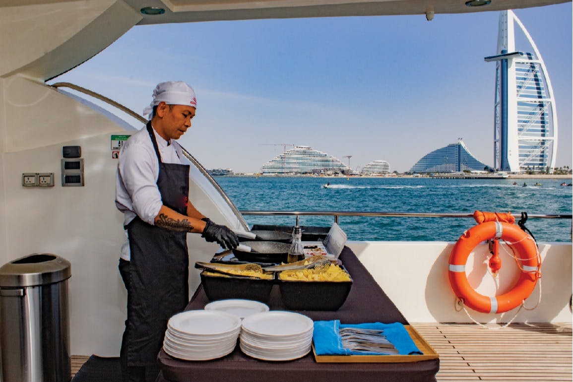 Kuchař v bílém klobouku a zástěře připravuje jídlo na palubě lodi s výhledem na oceán a moderní budovy v pozadí.