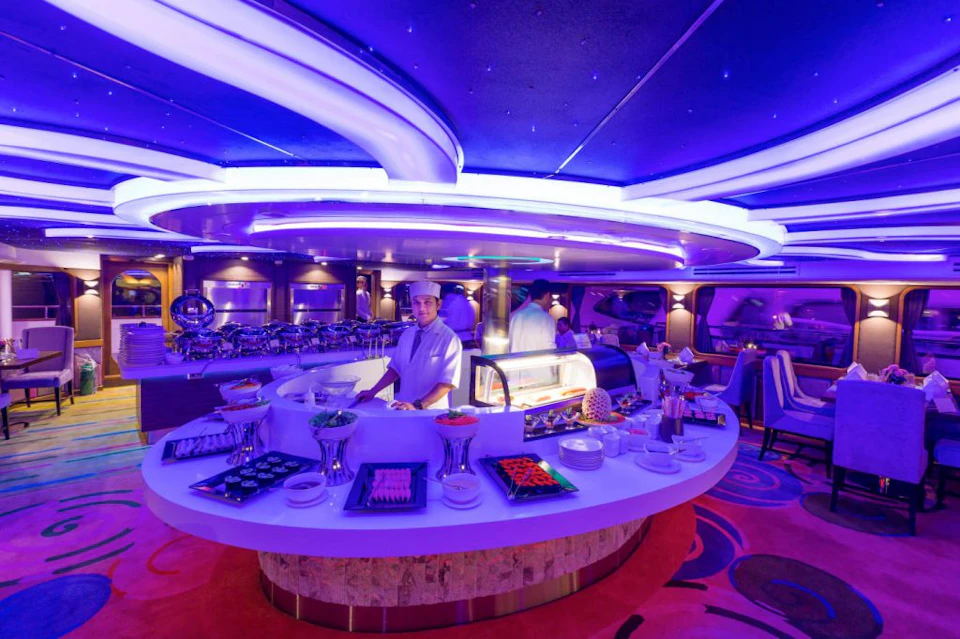 A chef stands behind a curved counter with various dishes in a modern, blue-lit buffet restaurant.