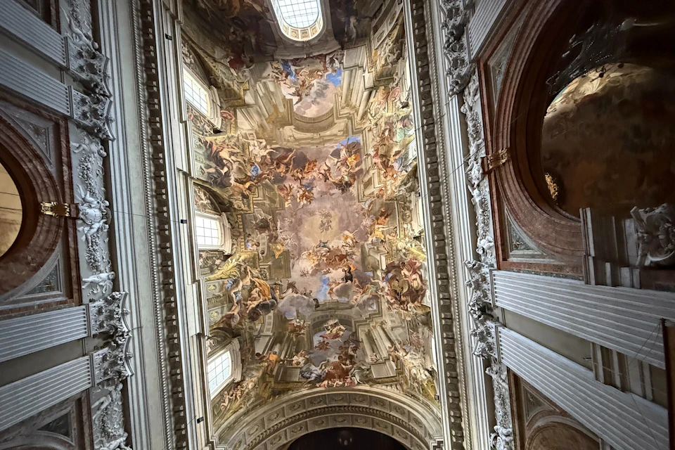 A richly decorated ceiling featuring elaborate frescoes and architectural details, with natural light coming through windows.