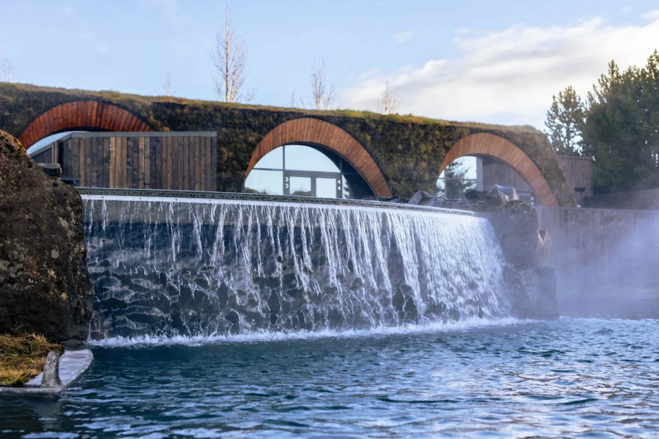 A modern building with arched windows and a green roof behind a cascading waterfall into a pool.