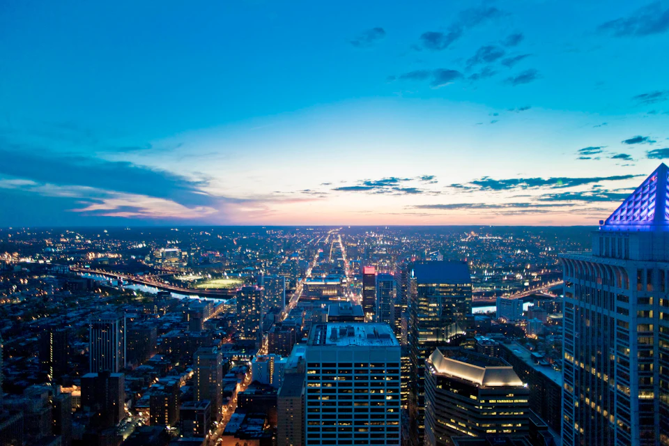Aerial view of a cityscape at sunset, with illuminated buildings, streets, and a river under a partly cloudy sky.